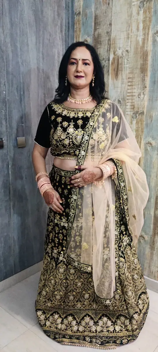 A woman in a black and gold embroidered lehenga with a sheer gold dupatta, henna on her hands, standing against a wall.