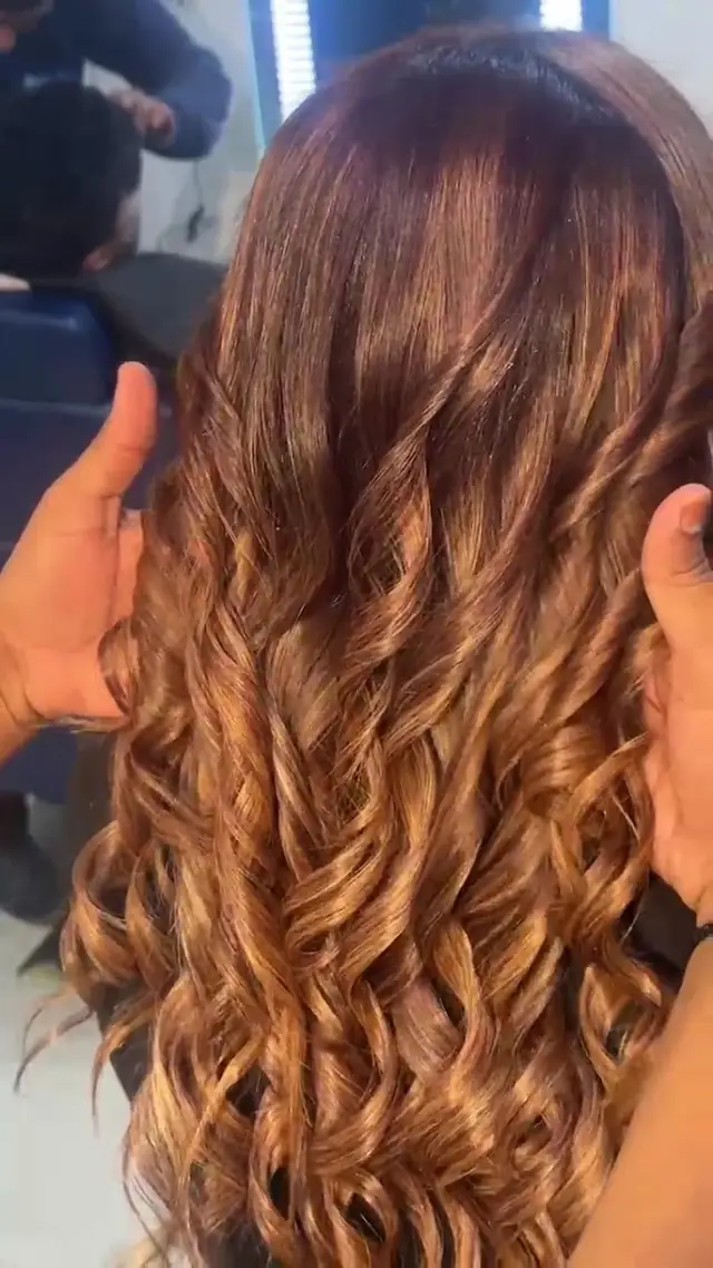 Long brown hair with caramel highlights being curled with a styling iron, seen from behind, two hands holding the curls.